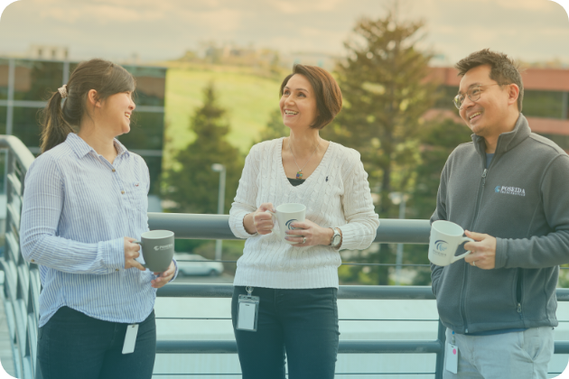 Three Poseida employees laugh while holding coffee mugs on an outdoor balcony.