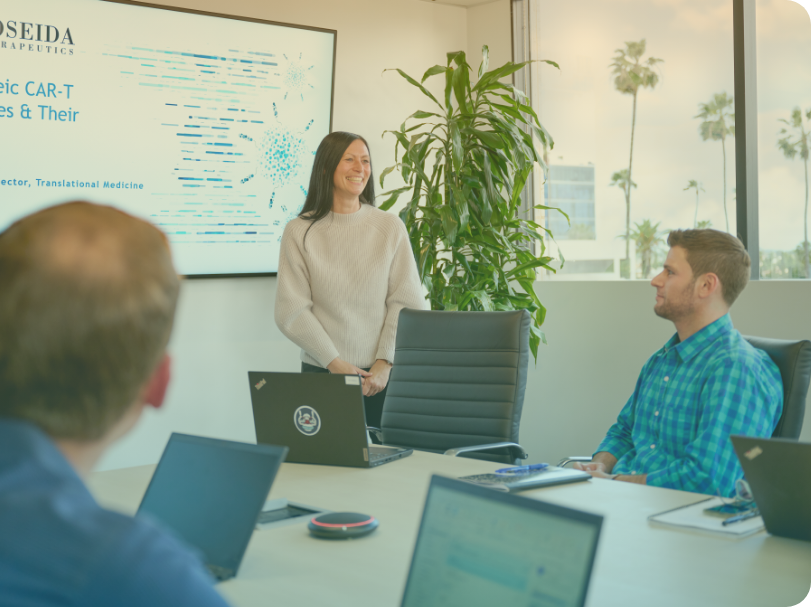 A Poseida employee stands at the front of a full conference room presenting slides displayed on a screen. Several employees sitting around the conference table look at the slides.